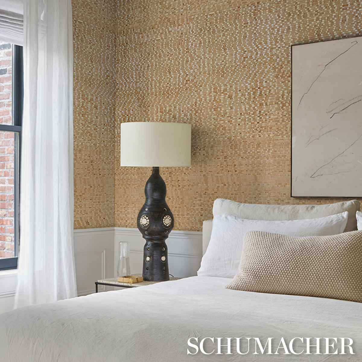 Bedroom with tan woven wallpaper, dark sculptural lamp, white bedding, beige pillows, abstract art, sheer curtains.