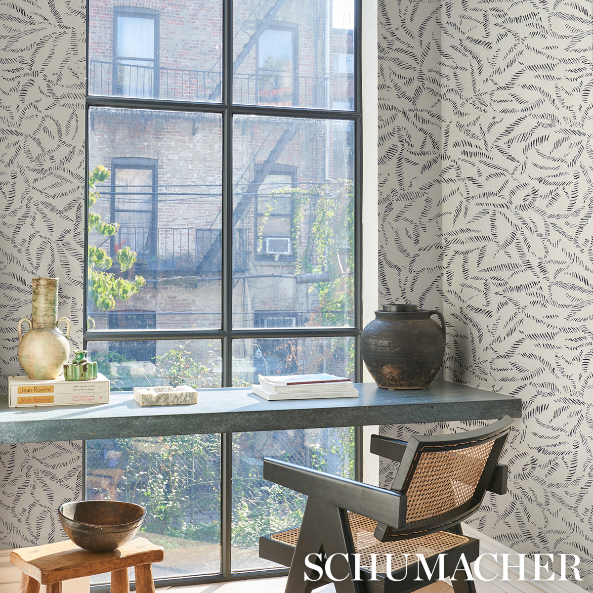 Loft-style workspace with large grid window, fern-pattern wallpaper, blue-gray desk, vintage vases, books, and wicker chair.