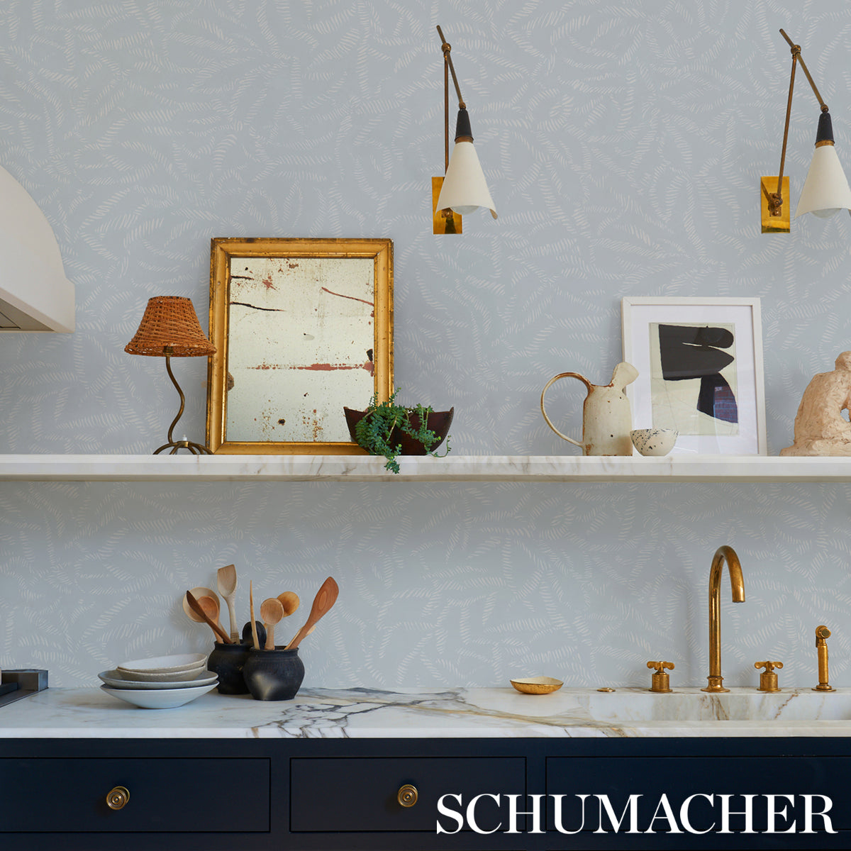 Marble kitchen countertop with navy cabinets, brass faucet, wall shelf holding framed art, lamp, and wooden spoons.