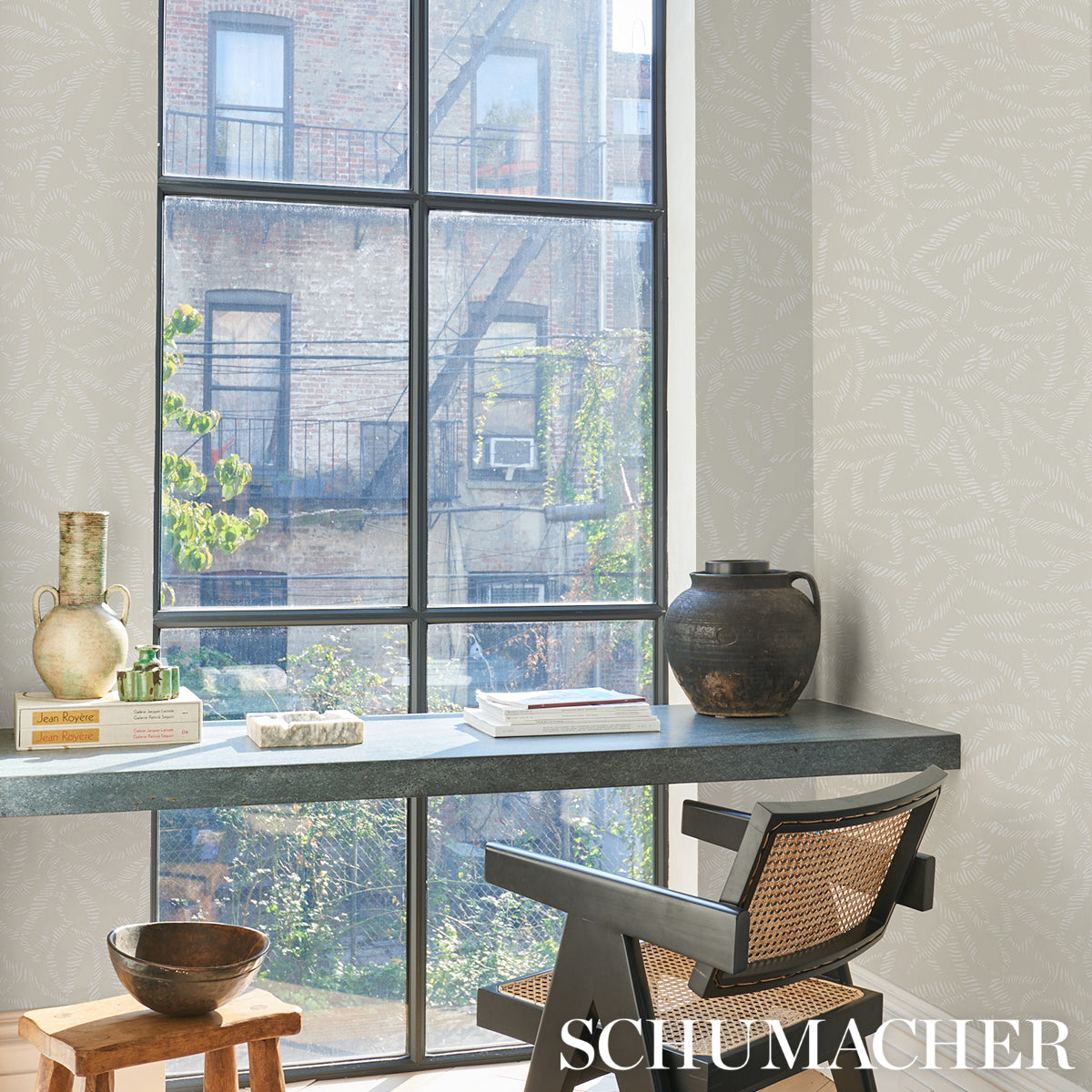 Industrial-style home office with blue-gray desk, black metal frame, rattan-backed chair, vase, books, and a large window.