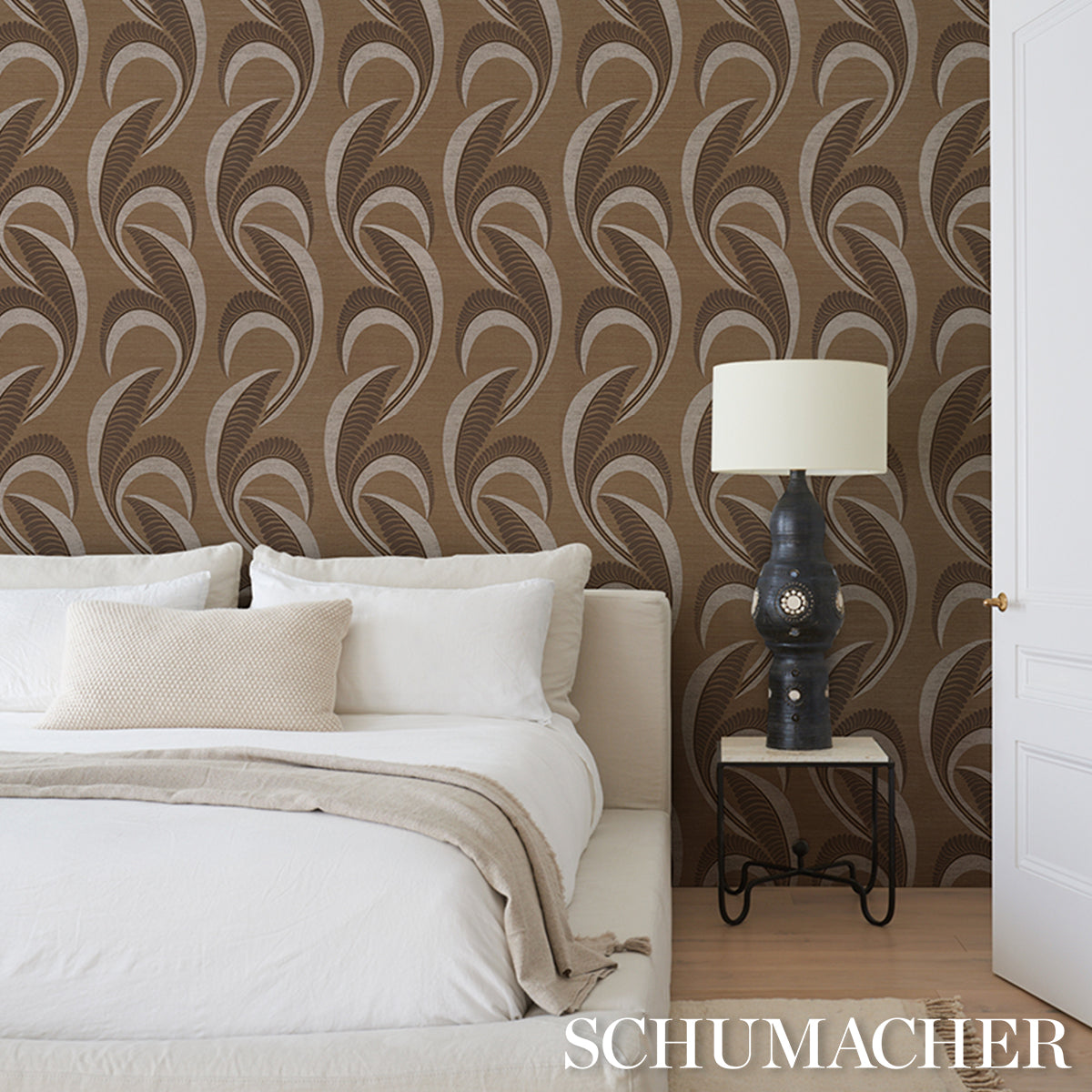 White bedding with beige throw, brown tropical leaf wallpaper, and a black curved-nightstand with a white lampshade.