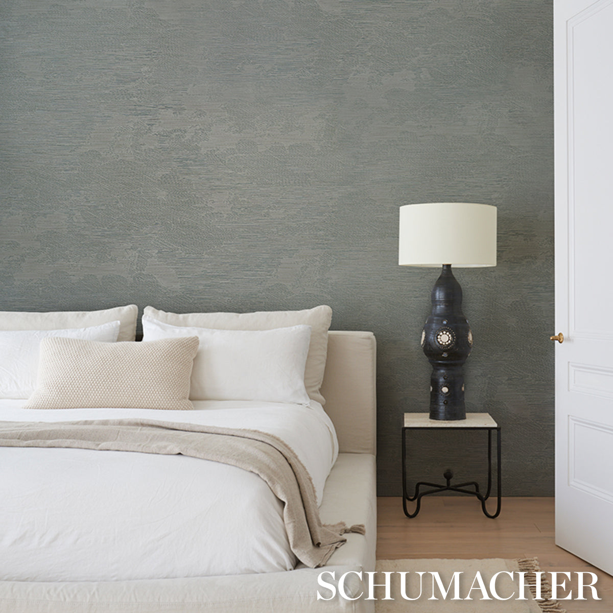 Bedroom scene with white bedding, beige throw and cushions; dark ceramic lamp on a metal nightstand beside gray textured wallcovering.