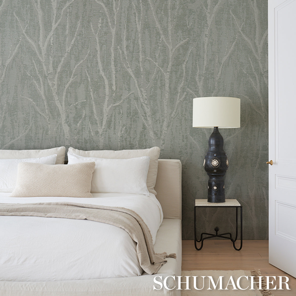 White bedding with beige throw and pillows, dark tree-pattern wallcovering, black lamp with cream shade on a metal nightstand