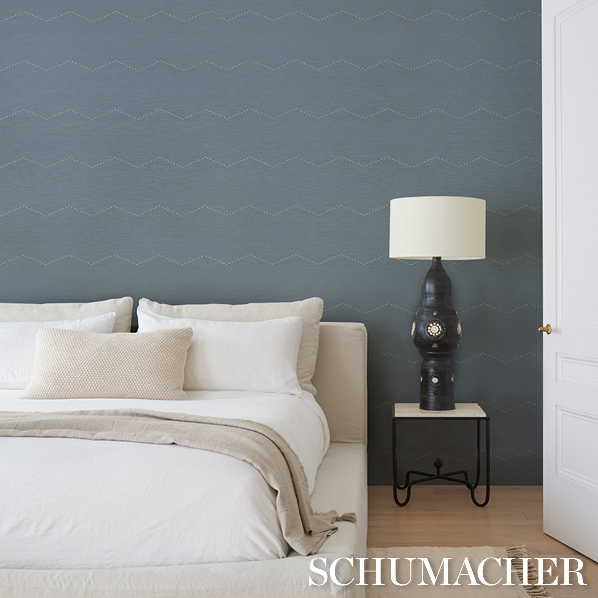 Dark blue wall with pale wavy pattern, white bedding with beige throw, black ceramic lamp on metal nightstand.
