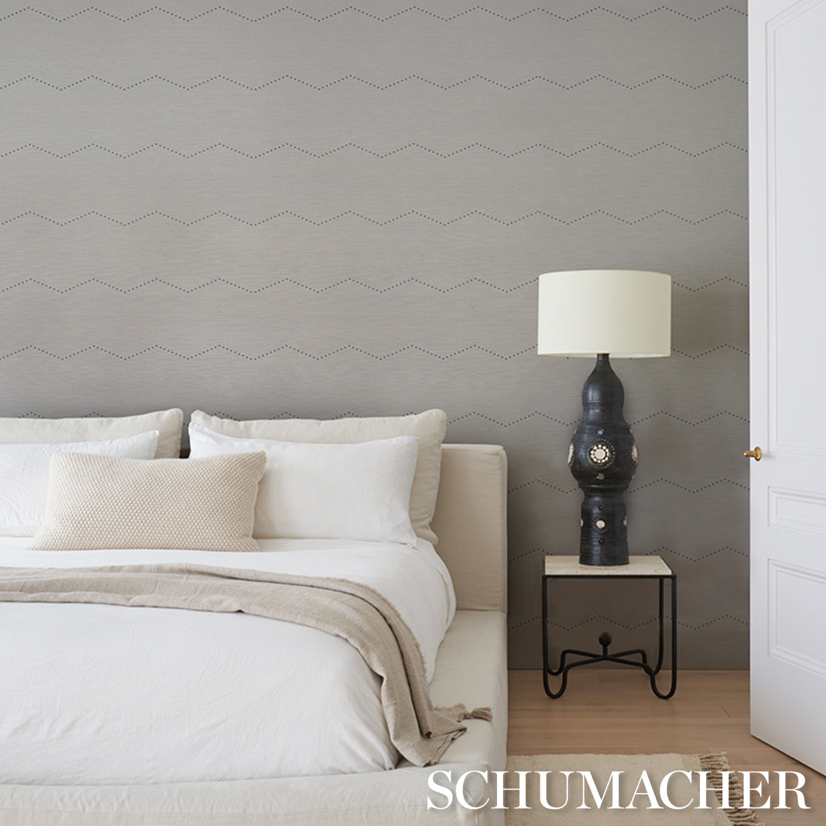 Neutral bedroom scene with white & beige bedding, taupe zigzag wallpaper, black ceramic lamp with cream shade on nightstand.