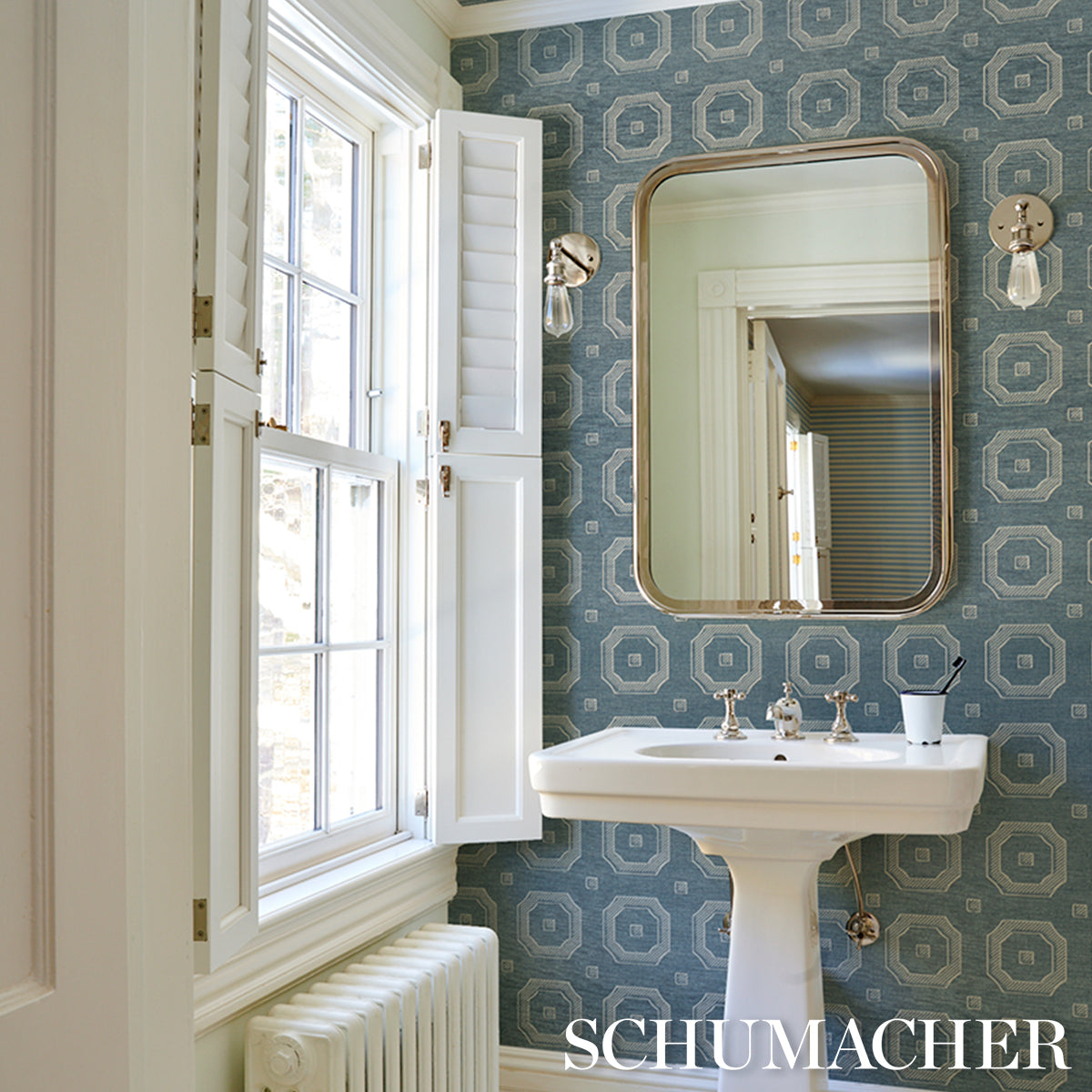 Blue geometric wallpaper bathroom with white pedestal sink, brass-framed rounded mirror, wall sconces, and window shutters.