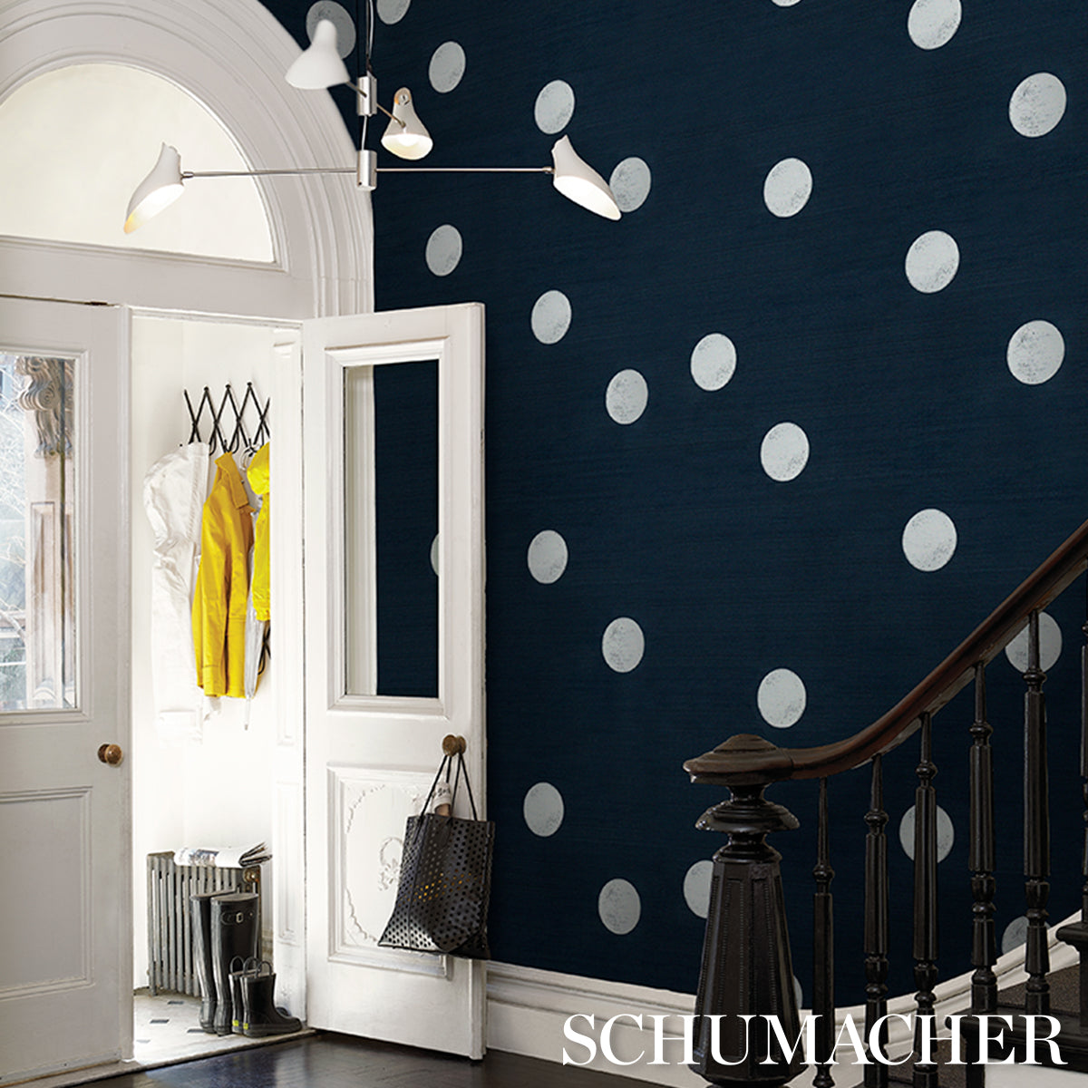 Entryway with navy polka-dot wallpaper, white doors, yellow coats in closet, dark wood staircase railing, chrome lamp.