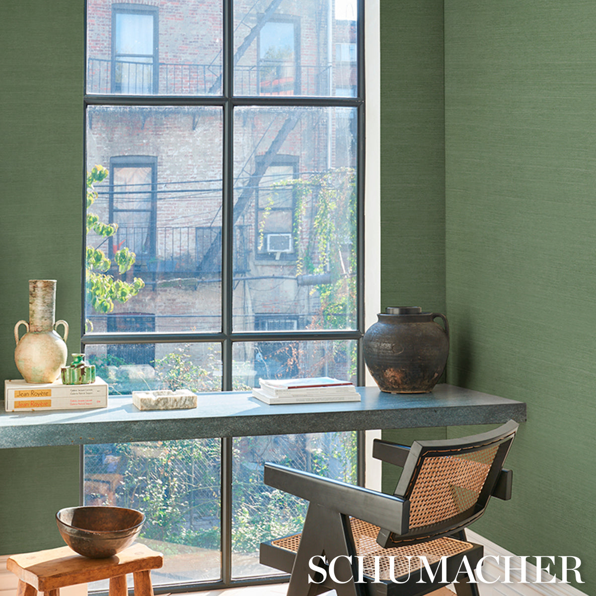 Green-walled sunlit home office; long gray stone-top desk, woven rattan chair, ceramic vases, books, clay pot by window.