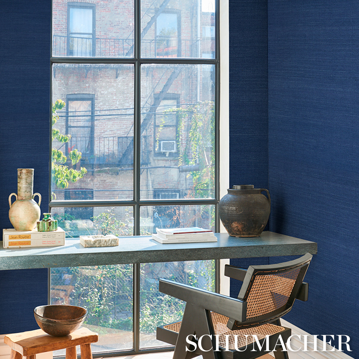 Blue-walled home office with a large window, gray stone desk, metal-framed woven chair, ceramic vase, and books.