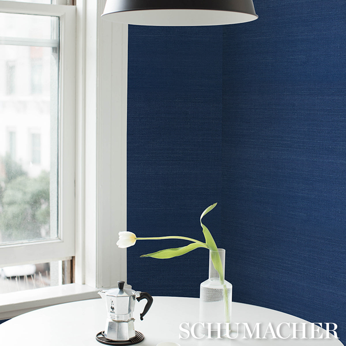 Blue textured backdrop with a white round table, an aluminum moka pot, and a glass vase with a white tulip.
