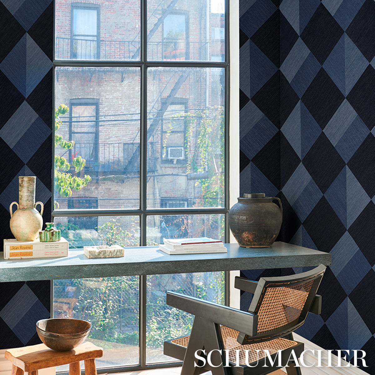 Bright room with blue diamond wallpaper, large window, stone table, cane-back chair, ceramic vase, bowls and books.