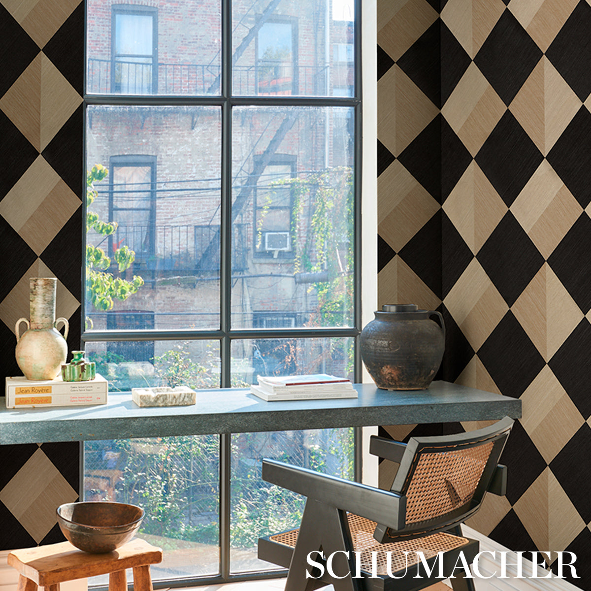 Light grey desk with books, a ceramic jug and bowl, black-and-beige diamond wallpaper, mid-century chair, urban window view.