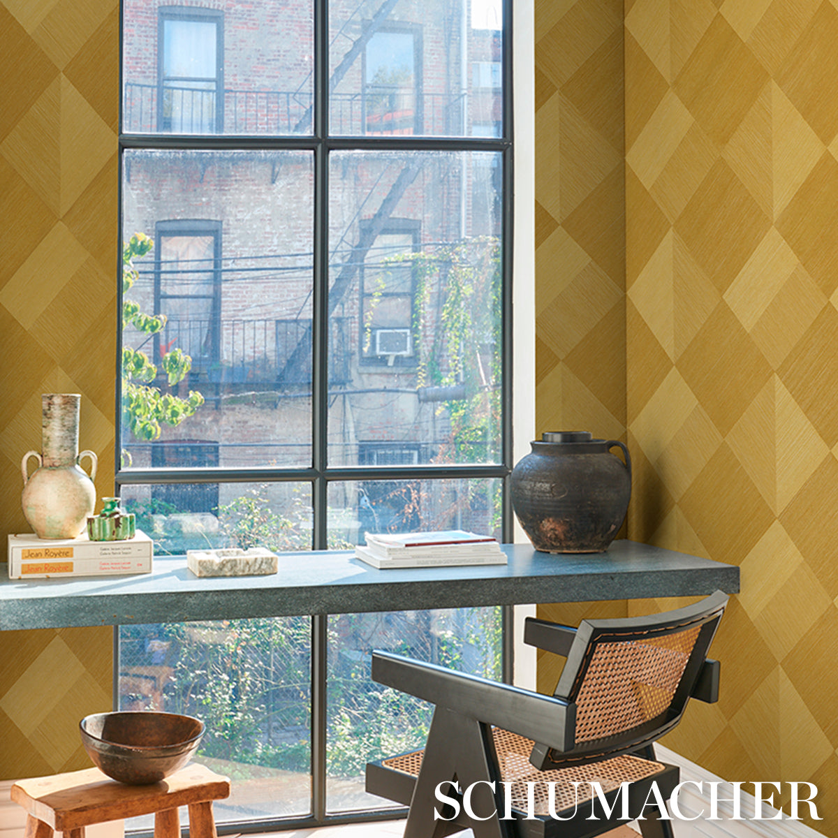 Sunlit window desk setup with yellow diamond wallpaper, gray metal desk, books, ceramic jug, woven cane chair, and dark pot.