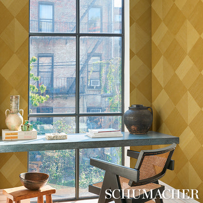 Sunlit window desk setup with yellow diamond wallpaper, gray metal desk, books, ceramic jug, woven cane chair, and dark pot.