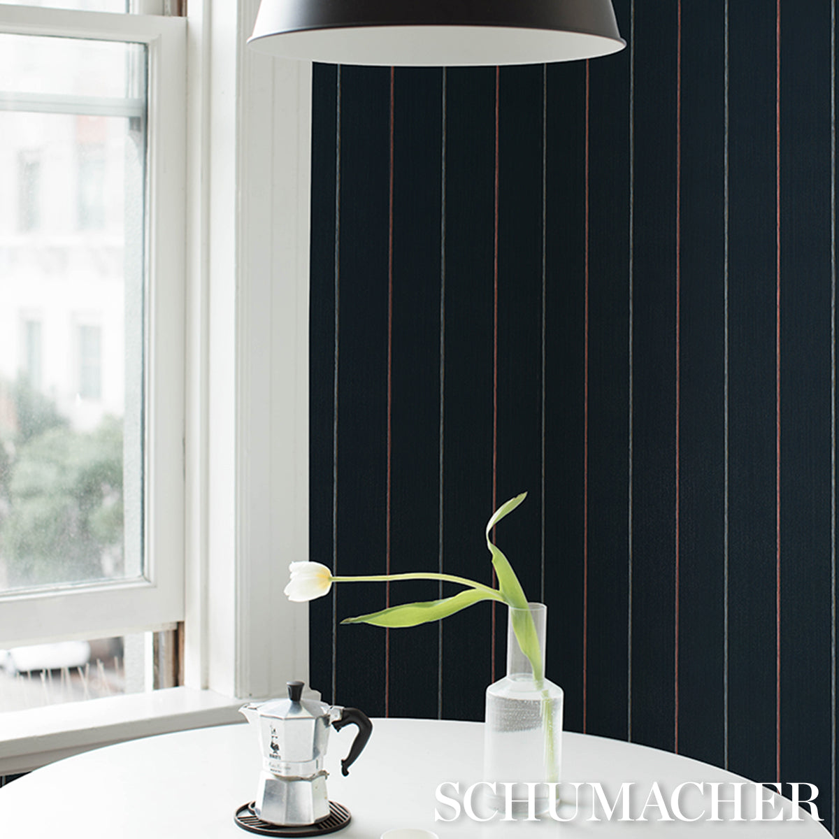 Dark navy striped wallpaper with thin red lines, white round table, moka pot, and tulip in glass vase under a pendant lamp.
