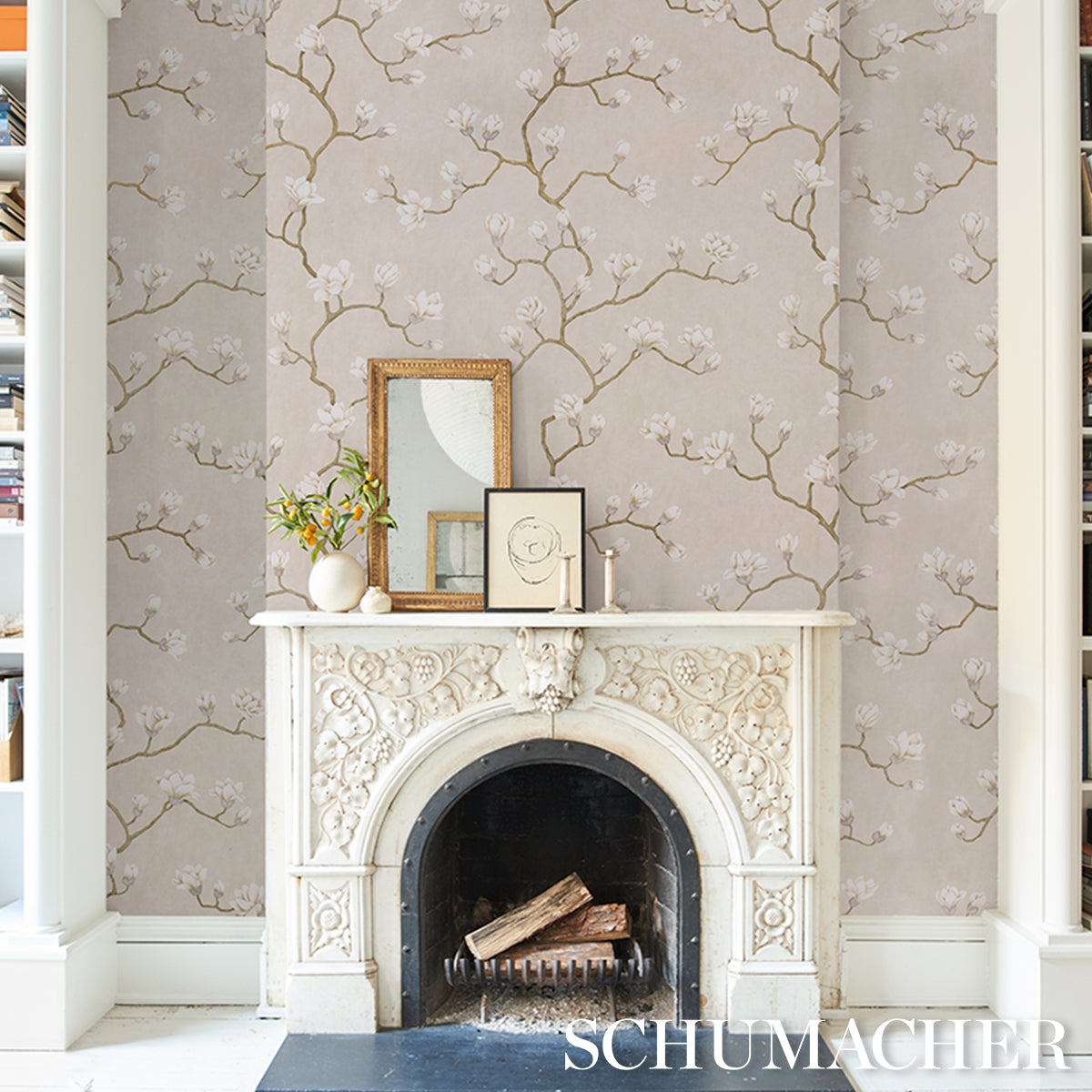 White ornate fireplace mantel with floral carvings and arched firebox, logs inside; beige wallpaper with blossom branches; framed art and vase atop.