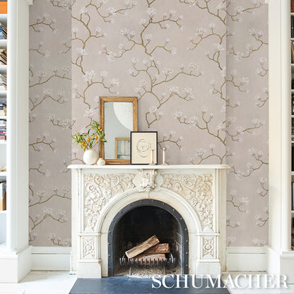 White ornate fireplace mantel with floral carvings and arched firebox, logs inside; beige wallpaper with blossom branches; framed art and vase atop.