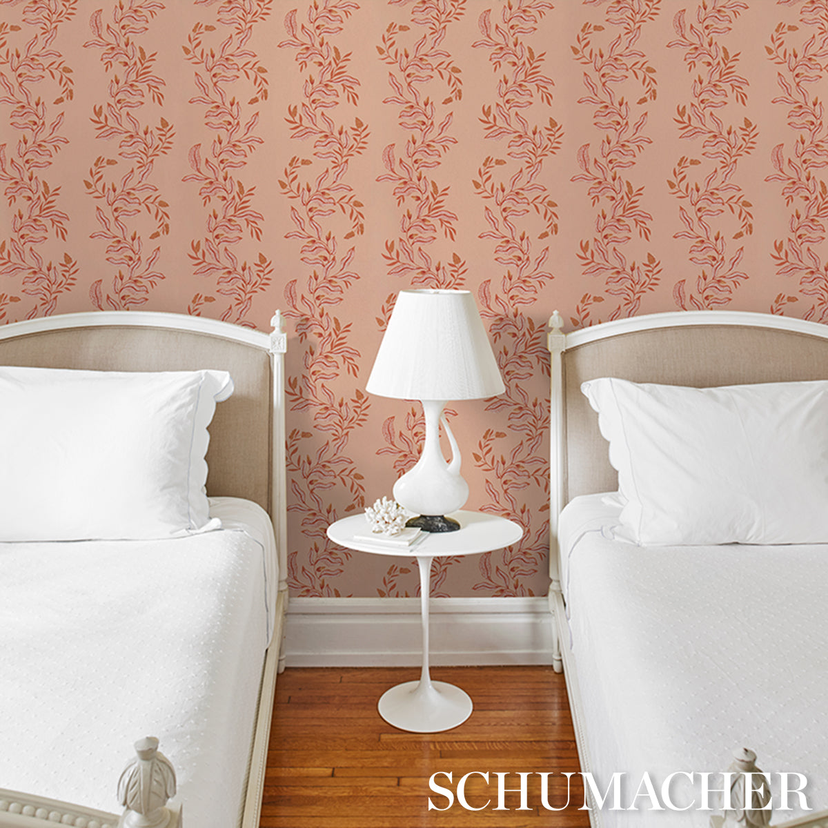 Twin white beds with beige headboards, pink floral wallpaper, white round nightstand with lamp between on wood floor.
