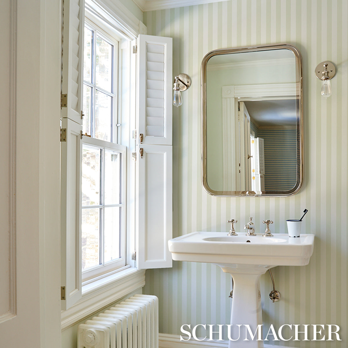 Pastel green striped wallpaper in a bathroom scene; white pedestal sink, brass-framed rounded mirror, wall sconces, window with shutters.