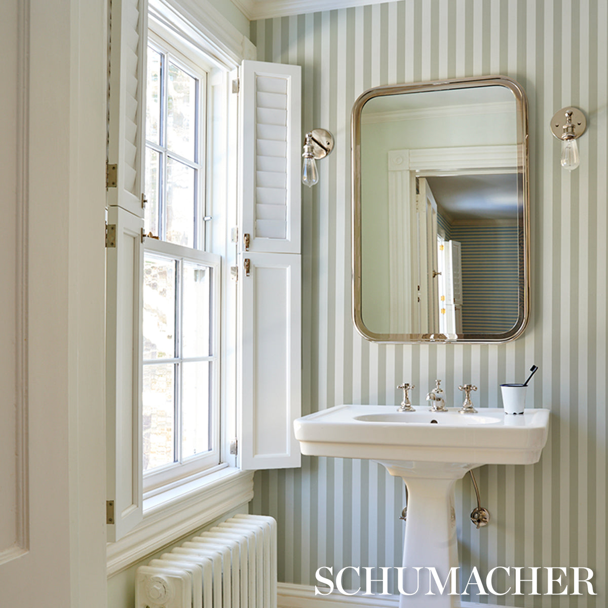 Vintage-style bathroom with pale green striped wallpaper, porcelain pedestal sink, brass-framed mirror.