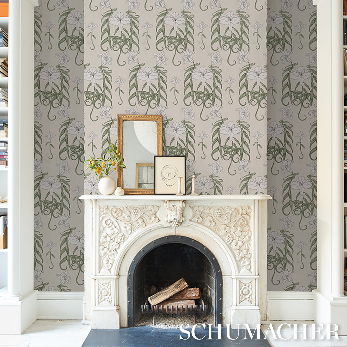 Ornate white carved fireplace with logs; grey-green damask wallpaper; mantel displays framed art & a vase.