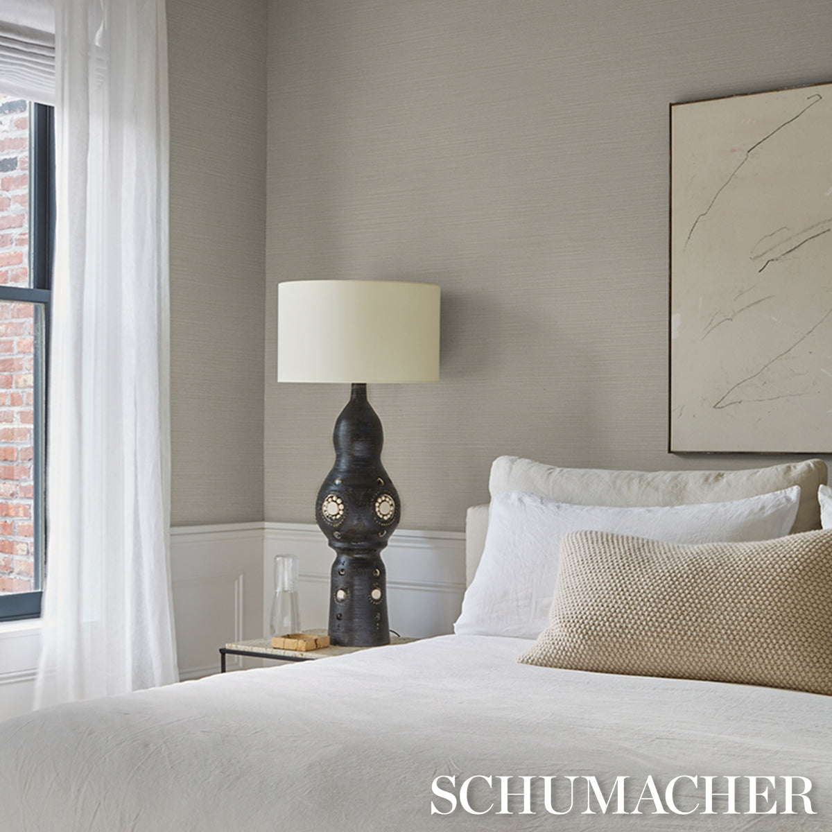 Neutral-toned bedroom with white bedding, beige accent pillow, sculptural dark lamp, sheer curtains, and limestone ABACA fine-weave vinyl wallpaper on the walls.