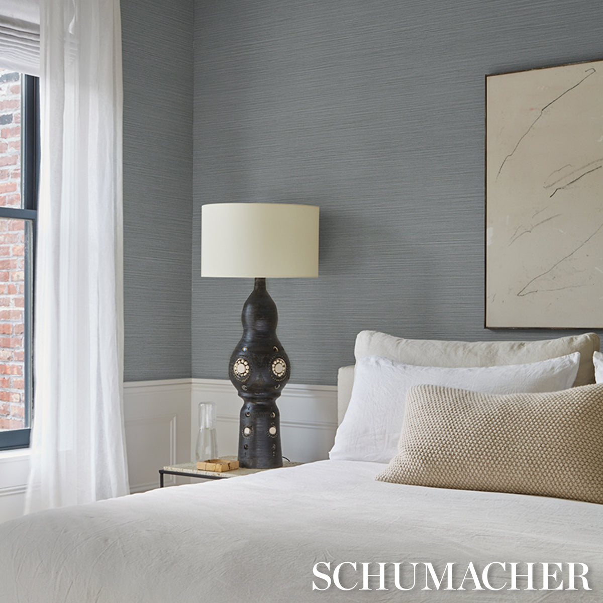 Bedroom interior with hazy blue-gray vinyl wallcovering, white bedding, beige throw pillow, dark sculptural lamp on nightstand, and sheer curtains.