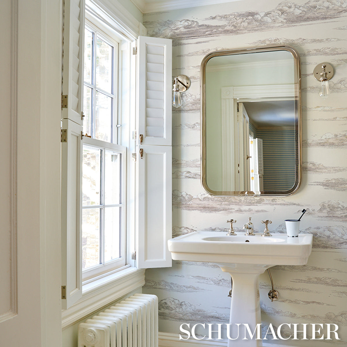 Bright vintage bathroom with pedestal sink, brass rounded mirror, white window shutters, striped wallpaper, toothbrush cup.