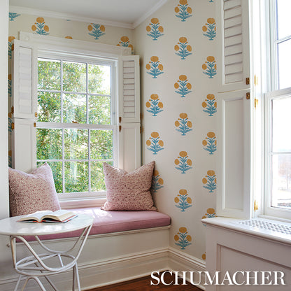 Pastel pink window seat with cushions, white shutters, blue-yellow floral wallpaper, and an open book on a white table.