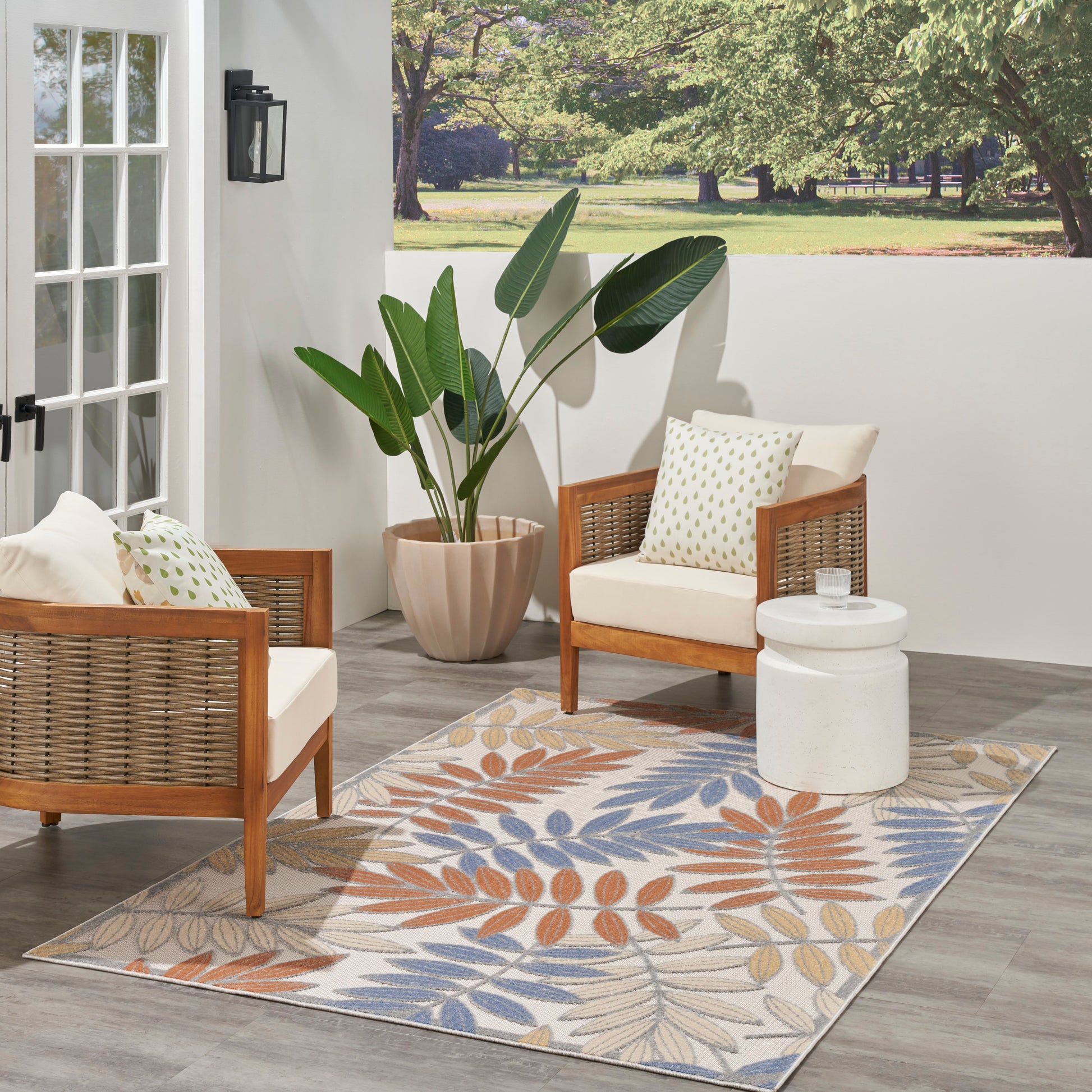 Outdoor patio with two wooden wicker armchairs, cream cushions, leaf-pattern rug, large potted plant, white side table.