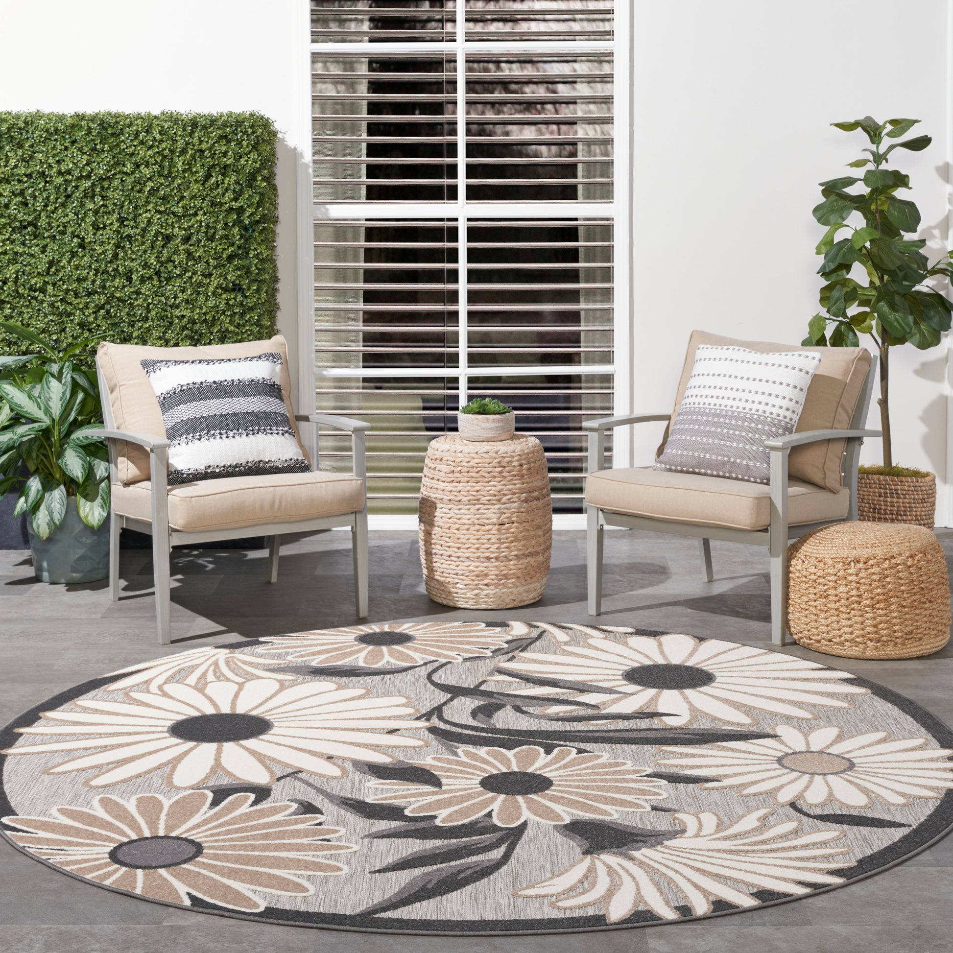 Outdoor patio seating: two beige cushioned chairs, round floral rug, woven side table, potted plants.