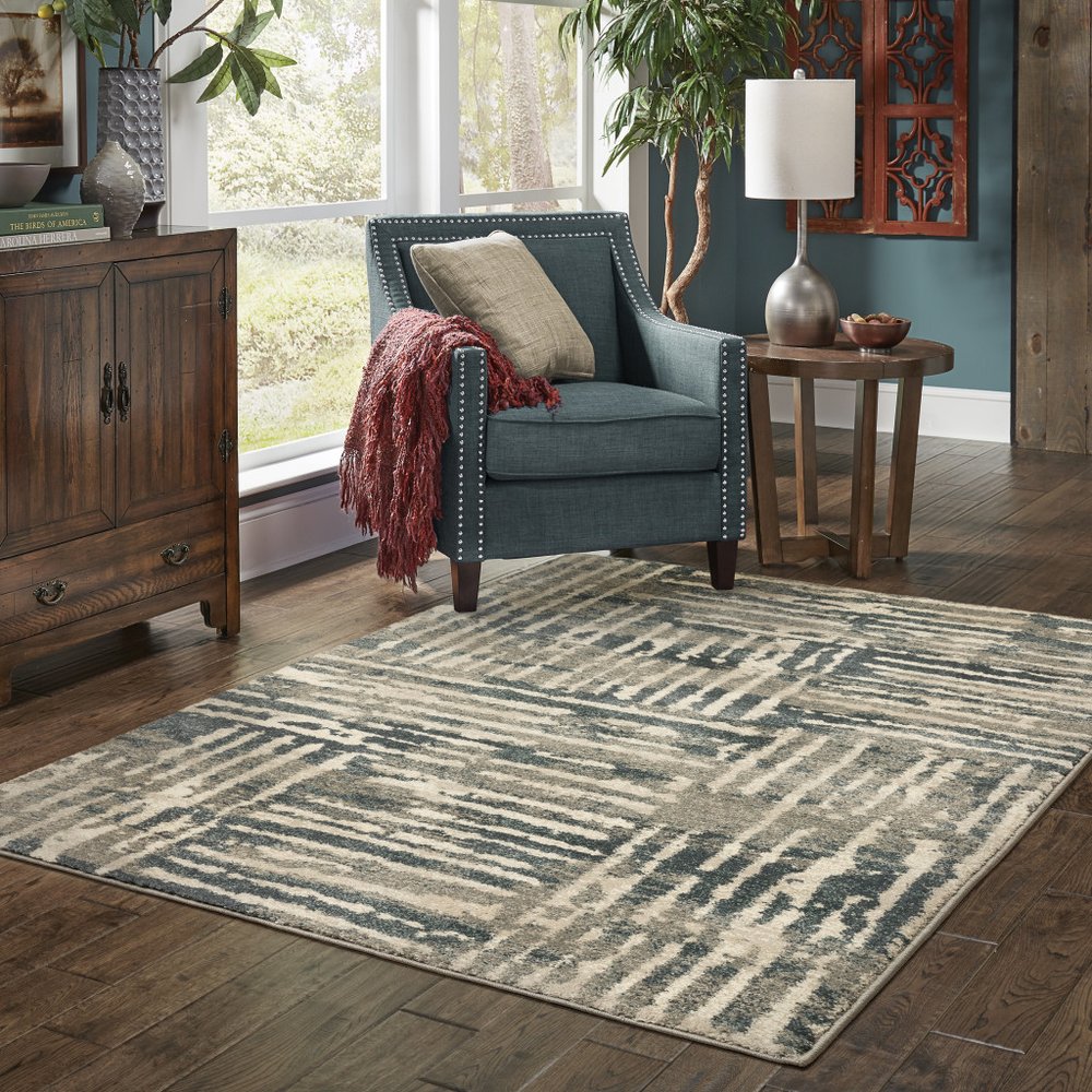 Blue upholstered accent chair with nailhead trim, red throw, wooden side table and lamp, on a gray geometric area rug.