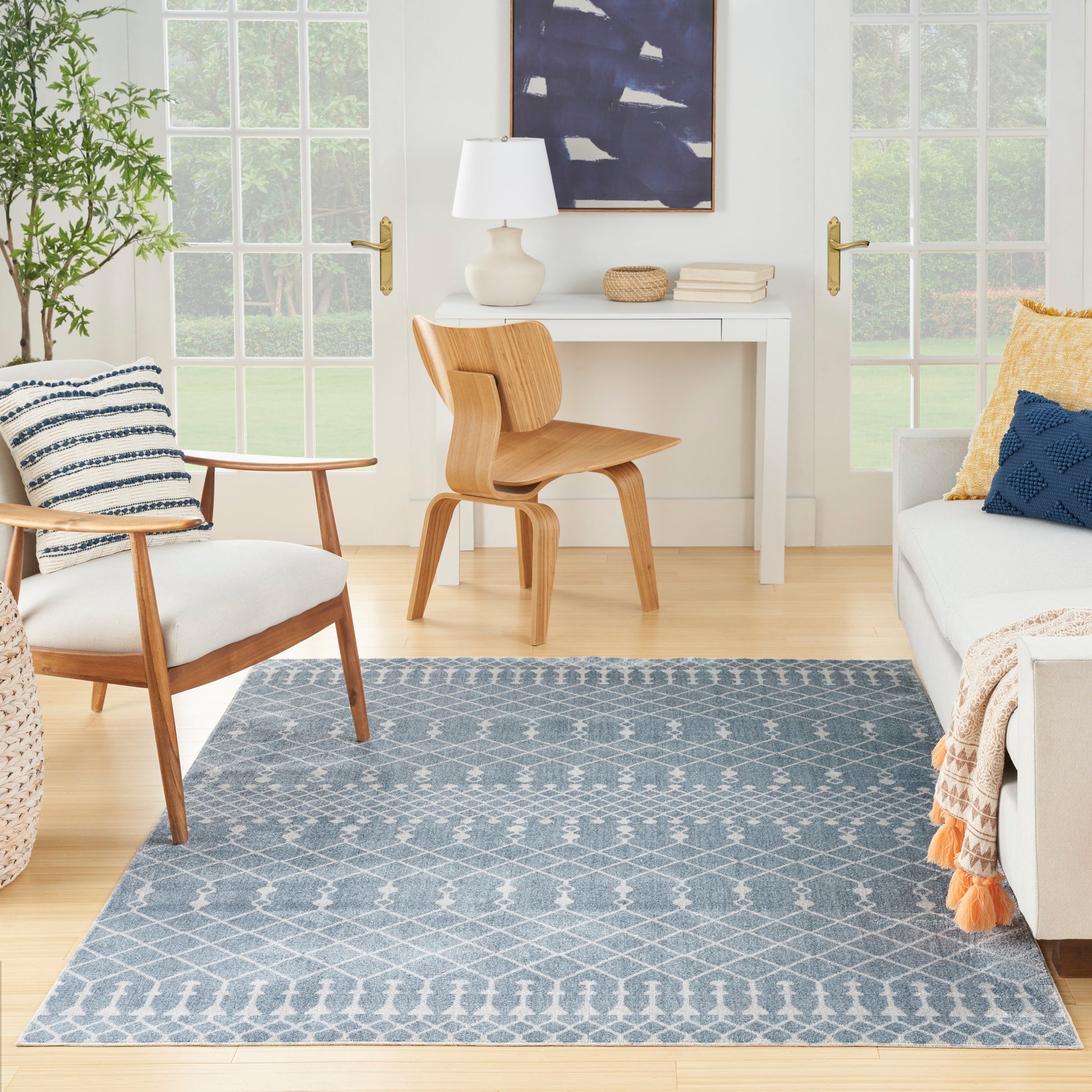 Bright living room featuring a blue geometric rug, mid-century wooden chairs, white desk, plant, and sofa with navy pillow.