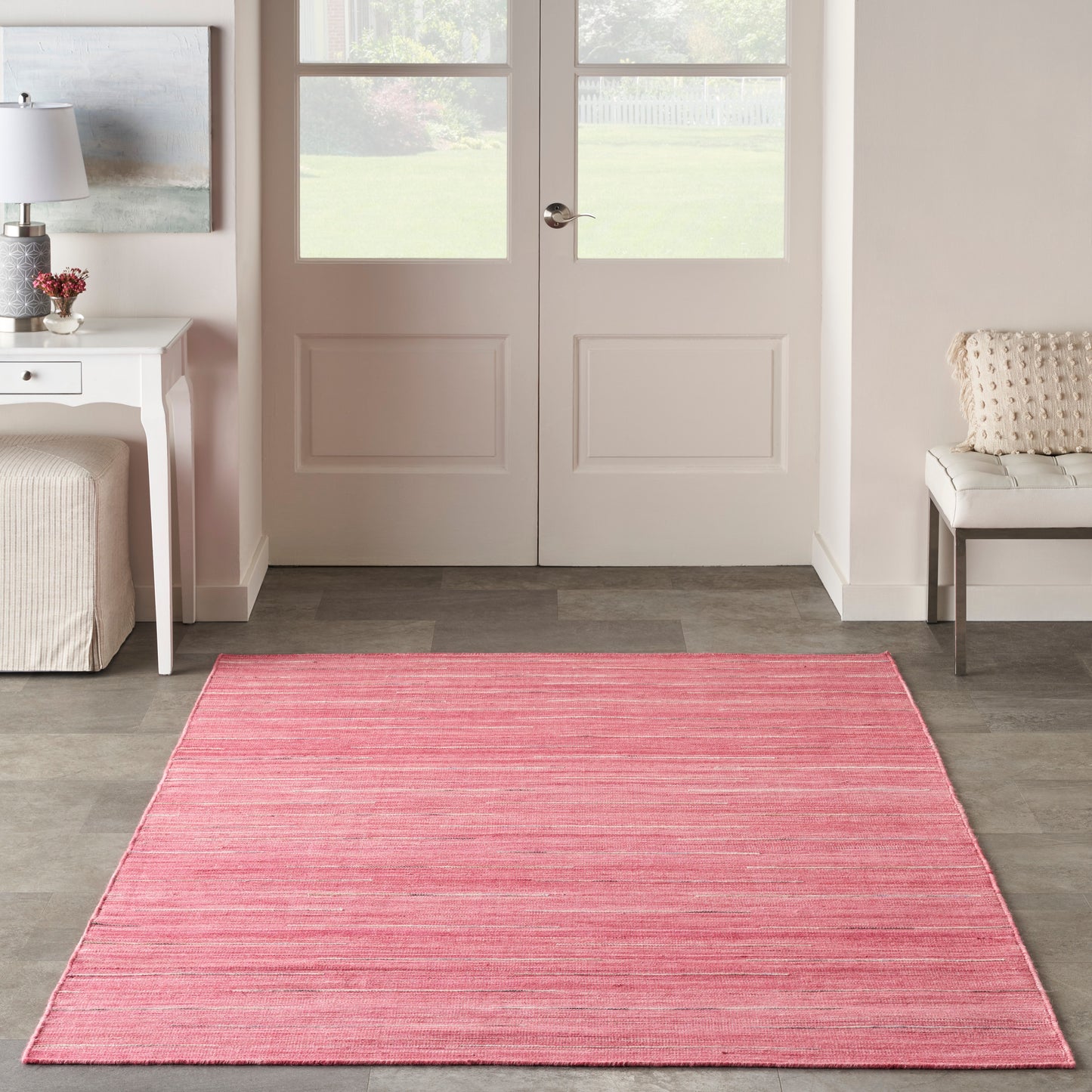 Pink rectangular flat-weave area rug with subtle tonal stripes in a bright entryway with white doors, side table and bench.