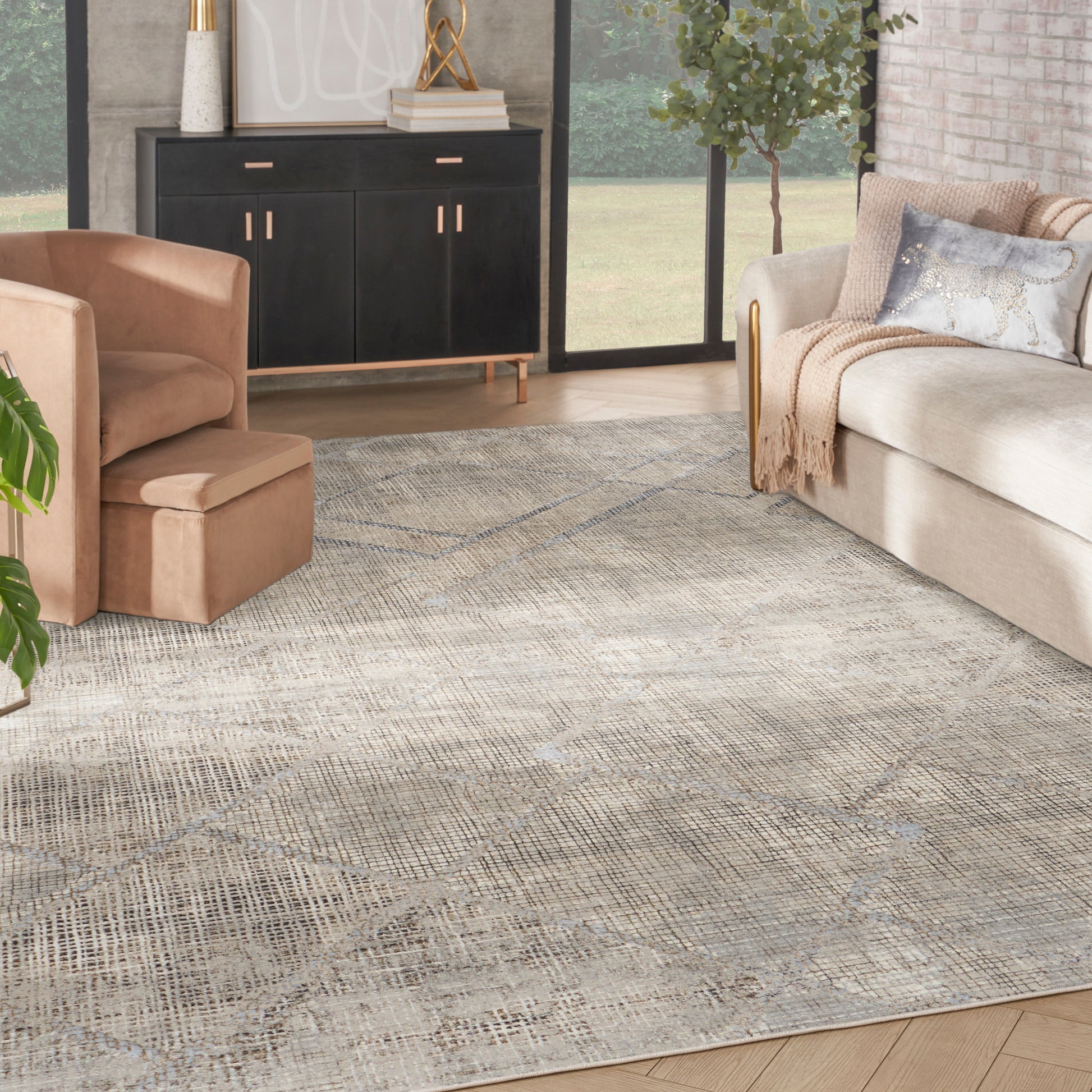 Large beige-gray flatweave rug with muted geometric lattice in a modern living room; tan sofa, peach ottoman, black cabinet.
