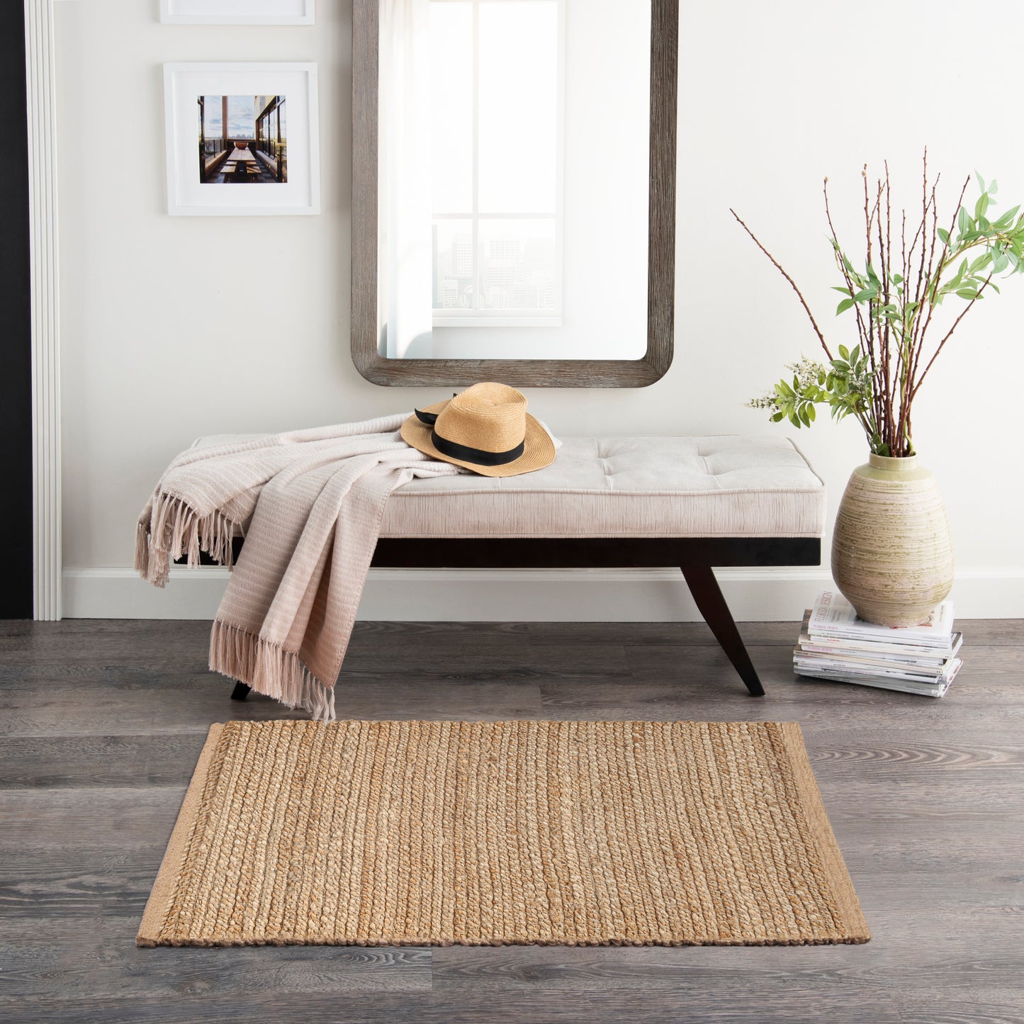 Neutral entryway with tufted white bench, beige throw, straw hat, wood-framed mirror, vase of branches, and jute rug.