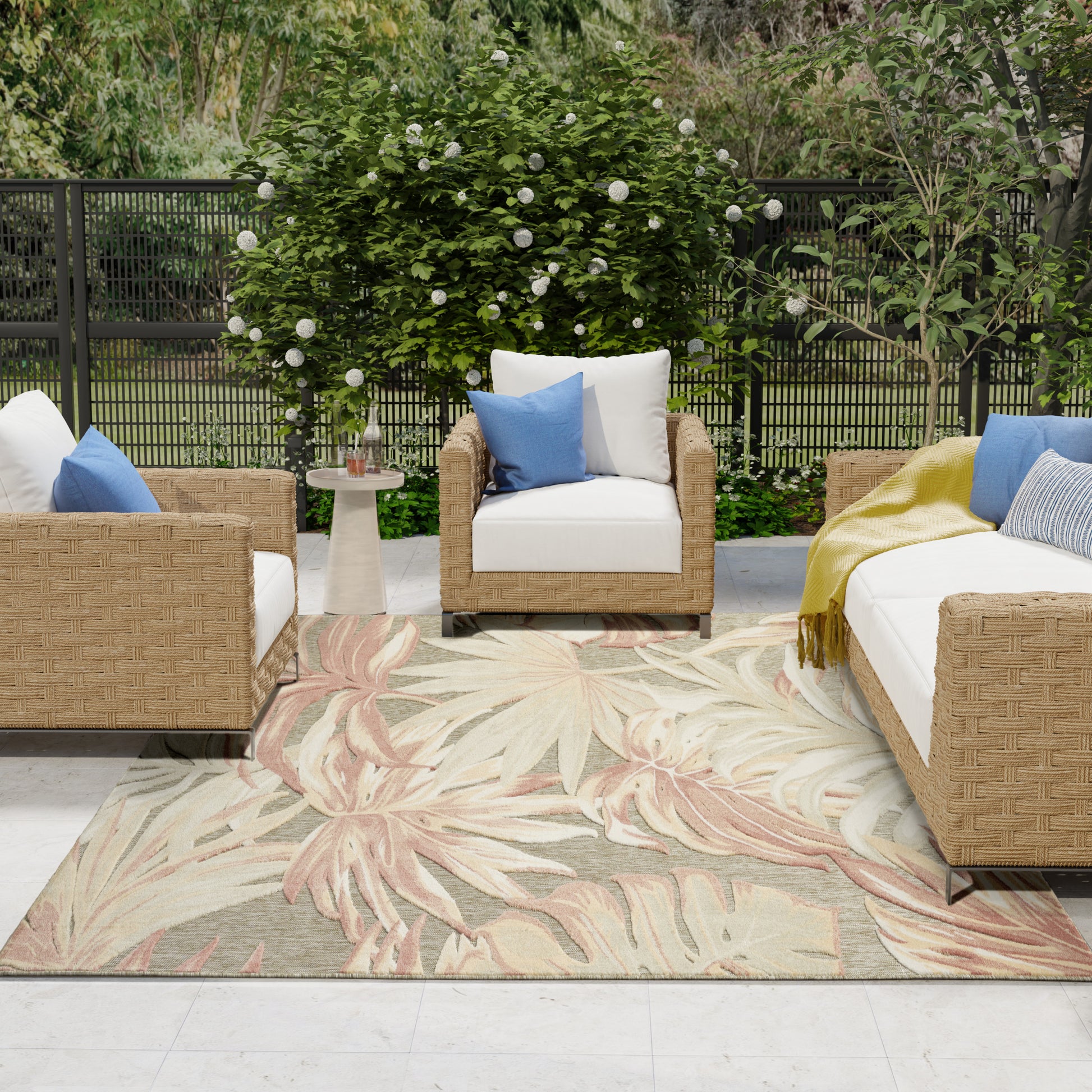 Outdoor wicker seating with white cushions, blue pillows, beige palm-pattern rug, and lush greenery backdrop.