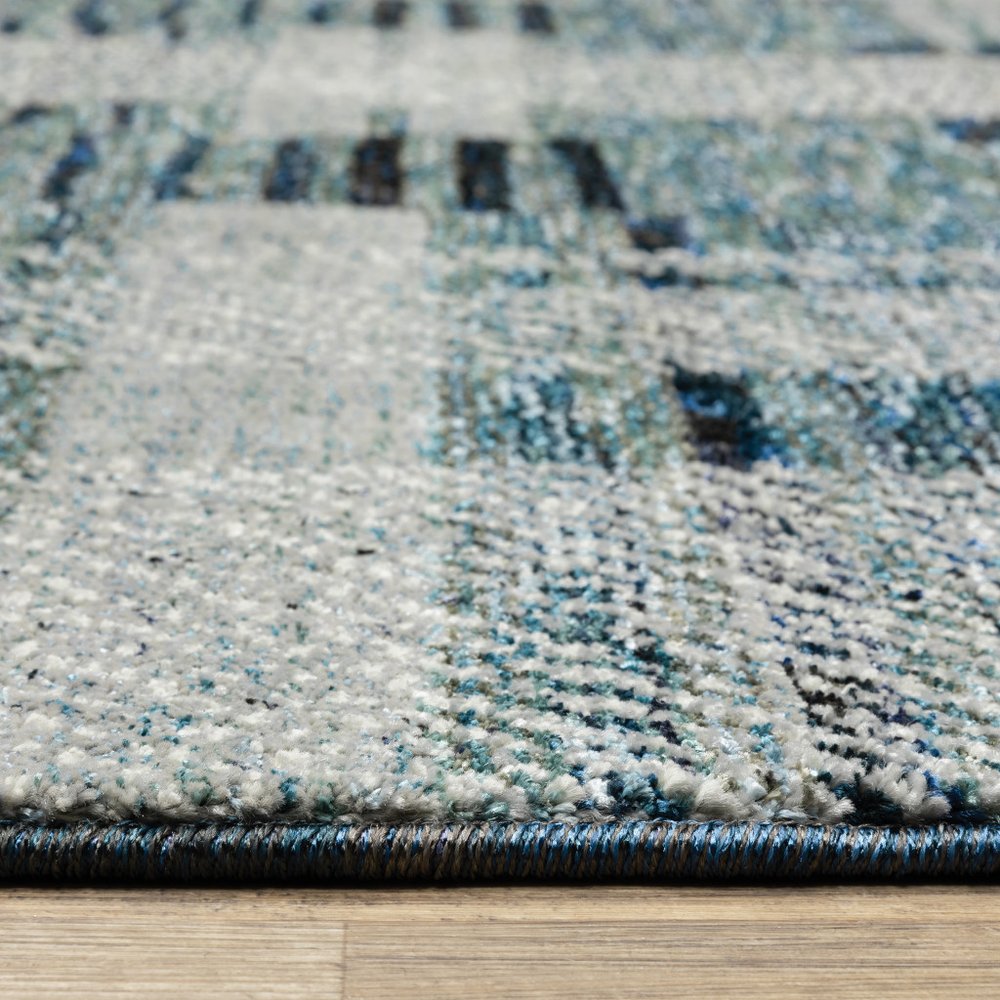 Close-up of a gray-blue patterned rug with woven texture and dark blue edging on a light wood floor.