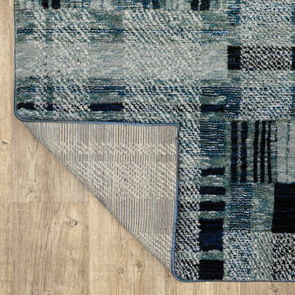 Blue-gray abstract area rug with geometric block pattern and blue edge binding; folded corner reveals light backing.