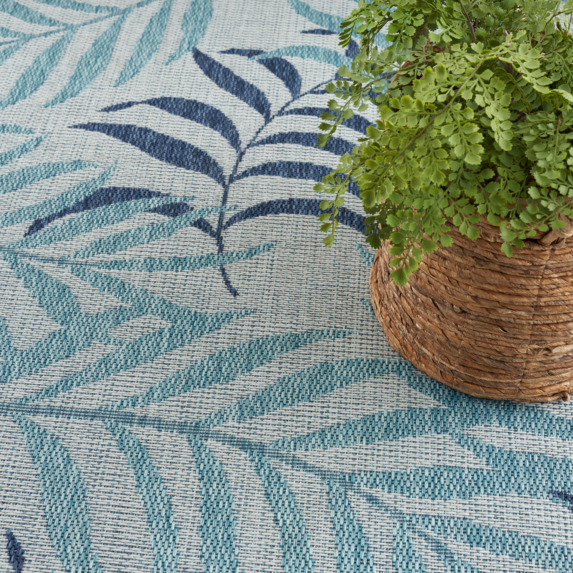 Teal and navy leaf-patterned woven rug with a potted fern in a wicker basket.