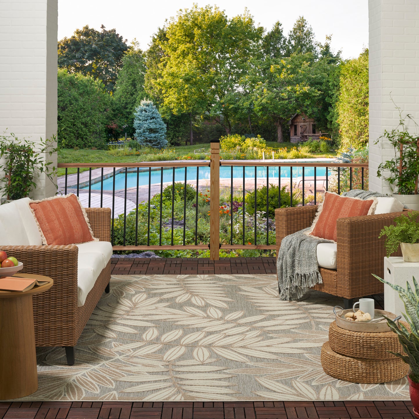 Leaf-pattern tropical rug on a covered patio floor with orange-cushion seating nearby, railing, garden and pool view, 7' x 10'.