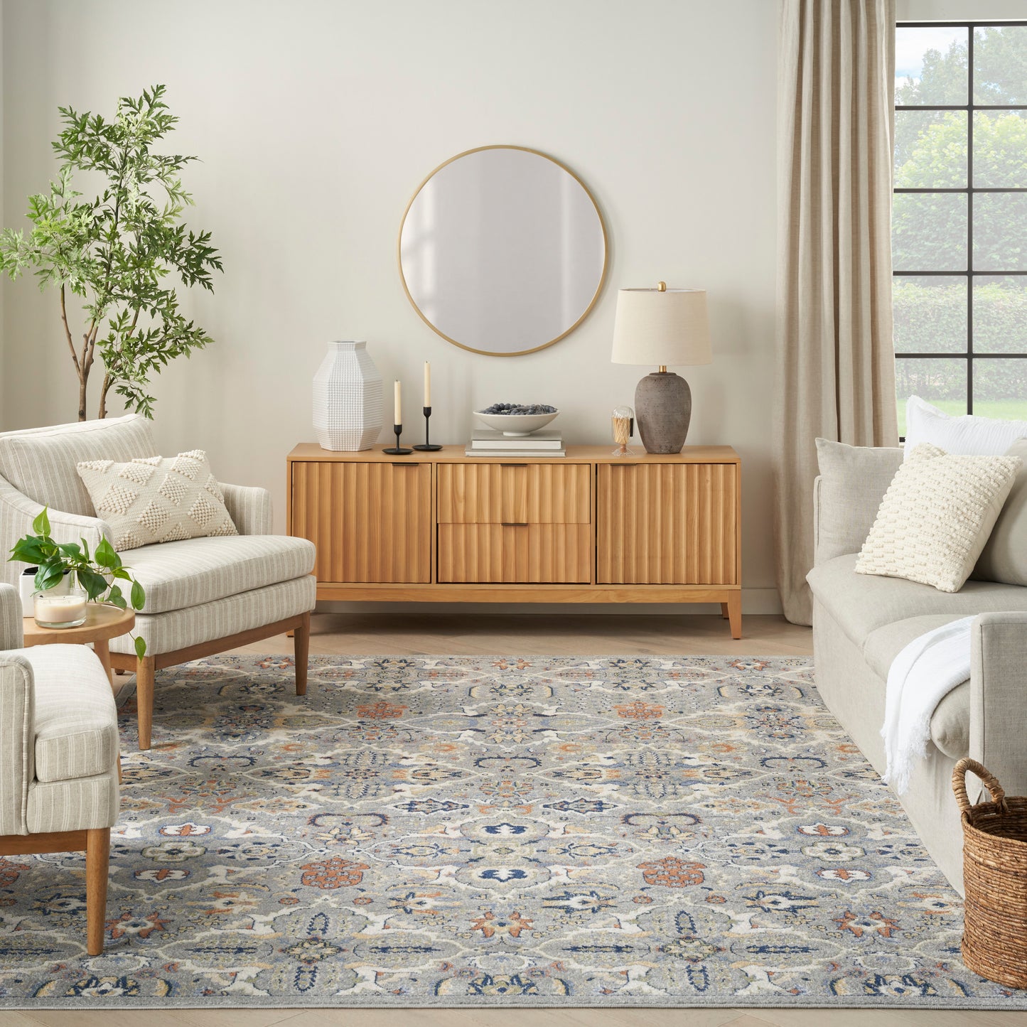 Mid-century teak sideboard with ribbed doors, round mirror, beige sofa and chairs, blue-gray patterned rug, greenery.