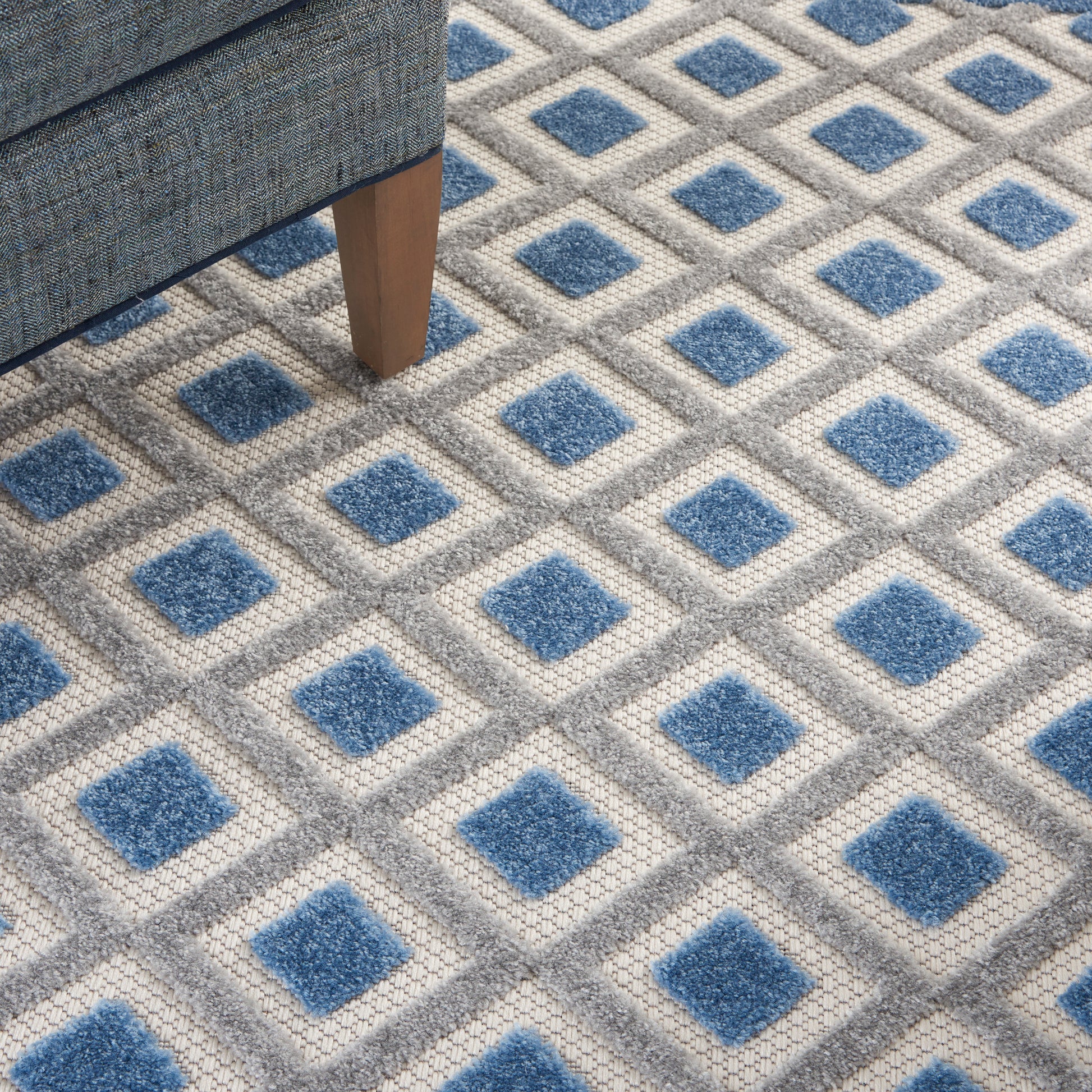 Close-up of a blue and gray diamond-pattern area rug; blue centers framed by light-gray diamonds, beside a wooden sofa leg.