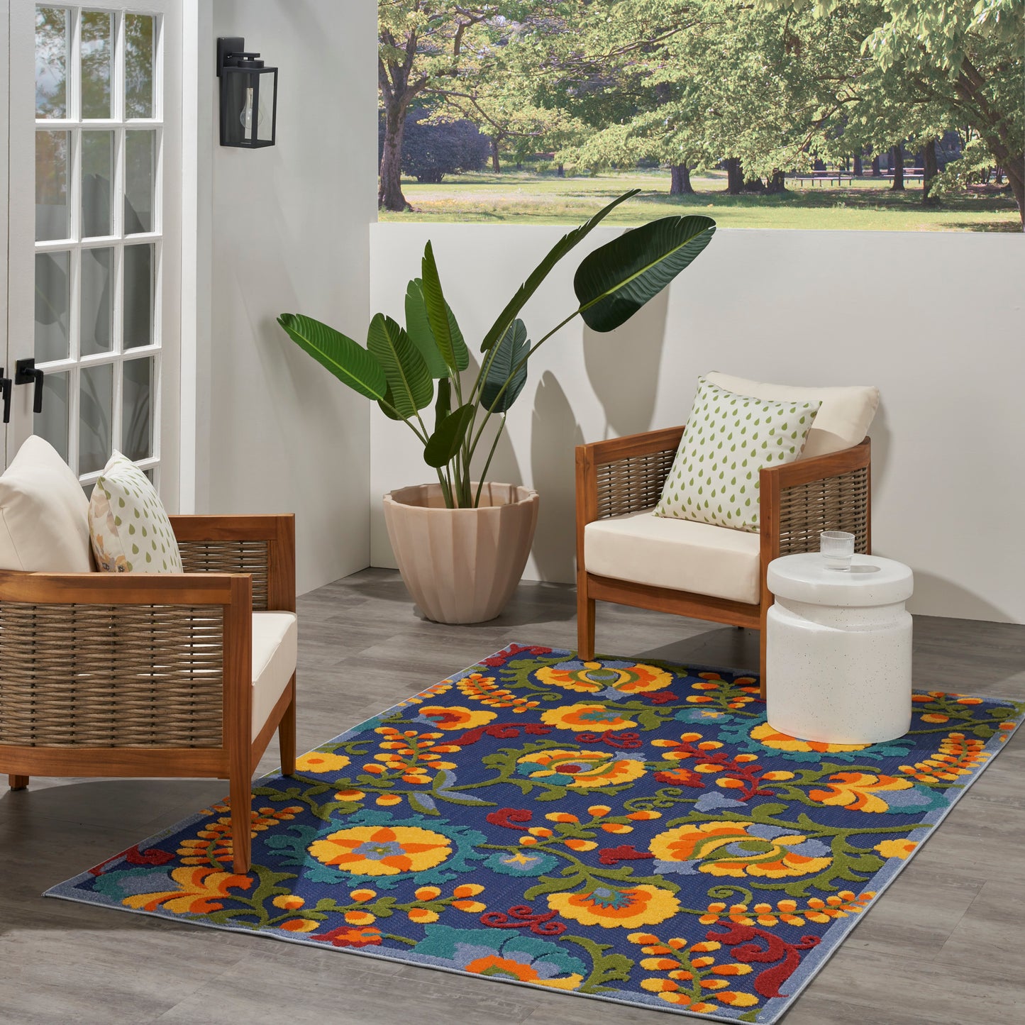 Outdoor patio with wicker-frame chairs, white cushions, colorful floral rug, large potted plant, and white side table.