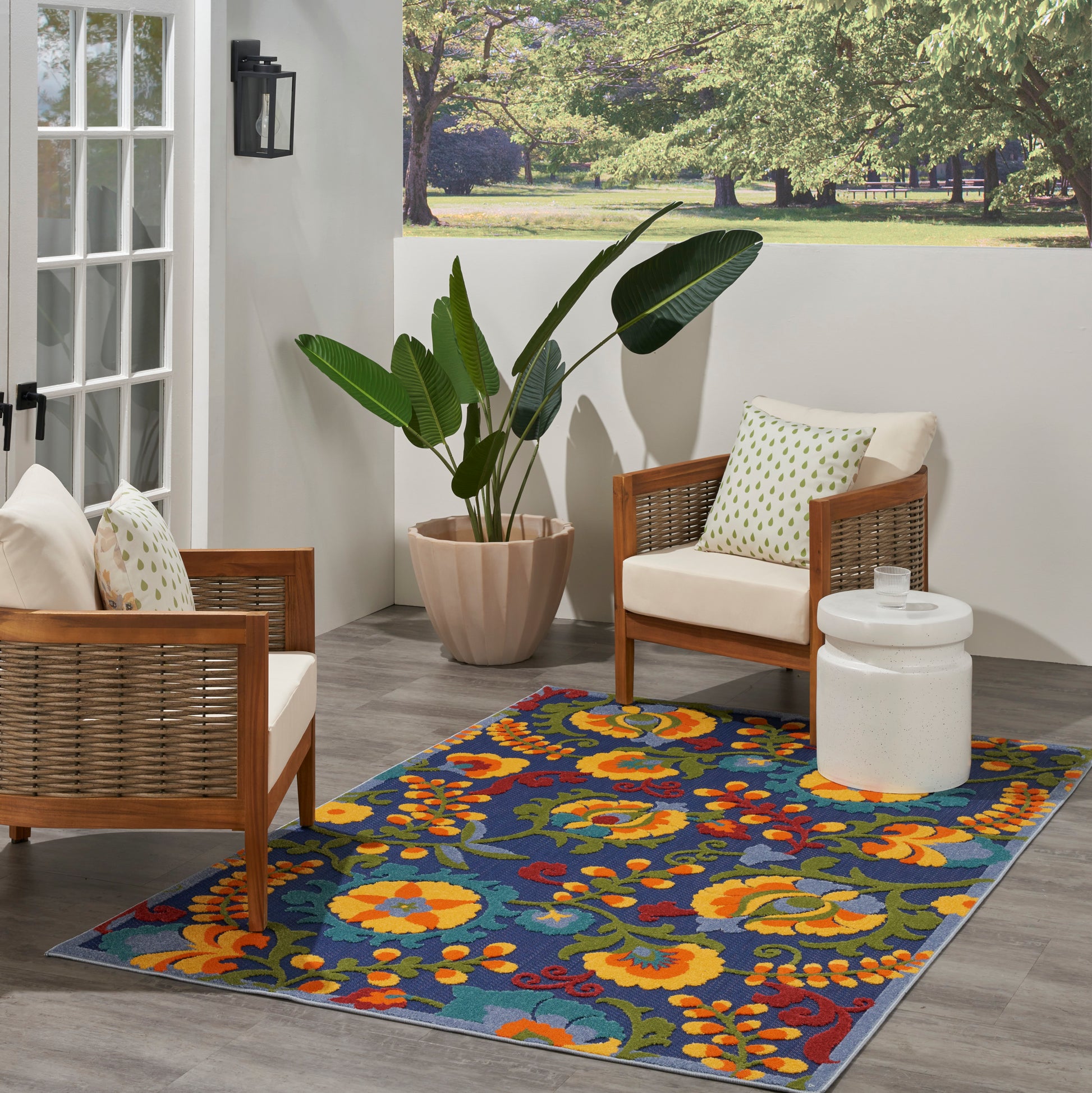 Outdoor patio with wicker-frame chairs, white cushions, colorful floral rug, large potted plant, and white side table.