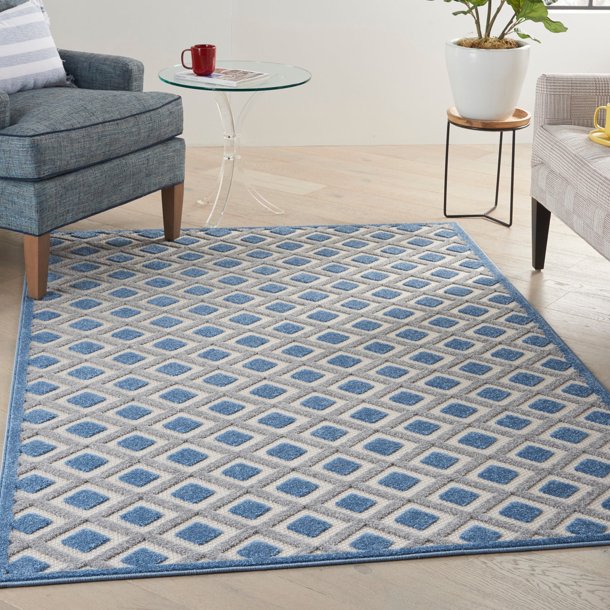 Blue diamond-patterned area rug in a bright living room with a glass coffee table, gray sofa, and potted plant.