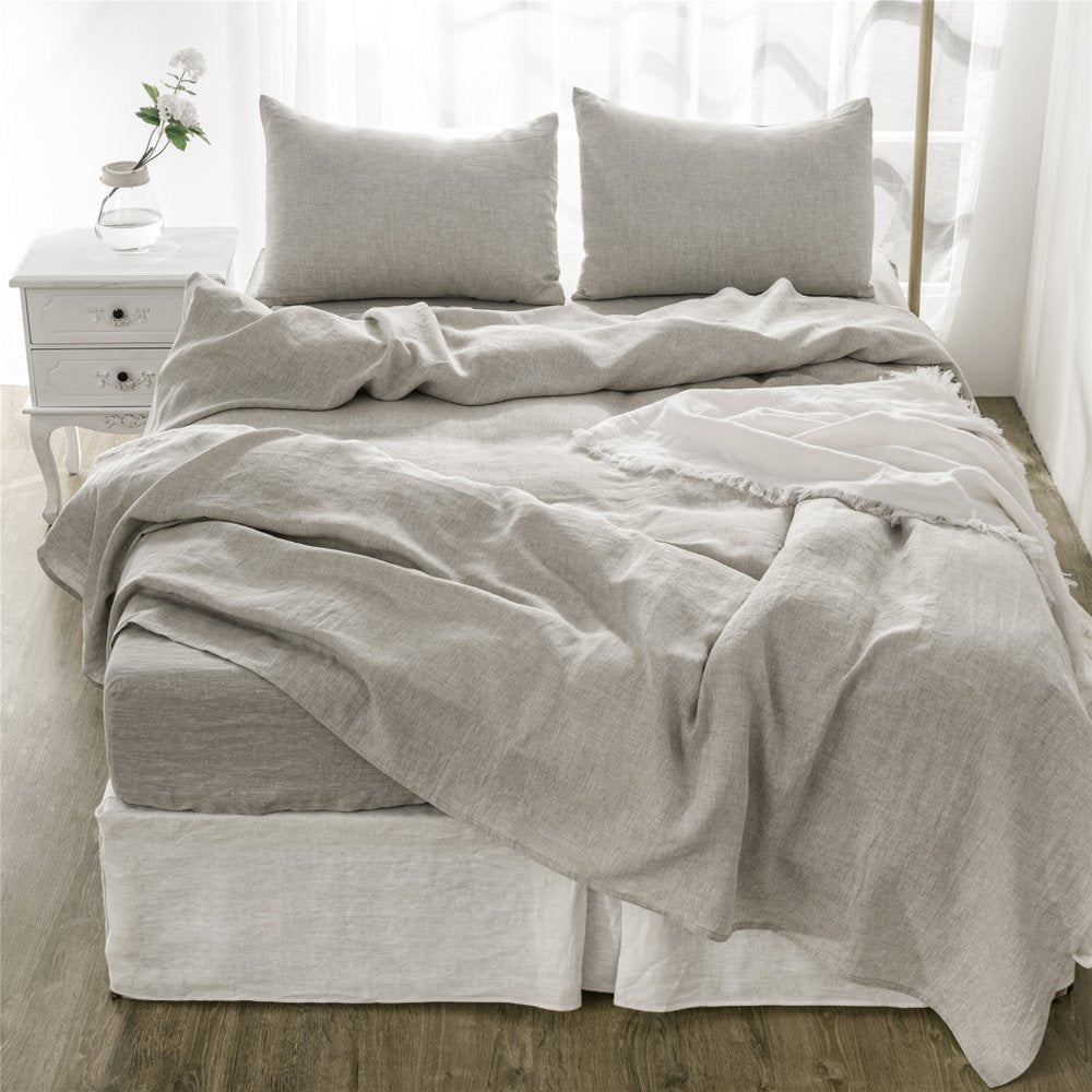 Gray linen bedding with two matching pillowcases on a neat bed in a bright, airy room & white nightstand with a vase.