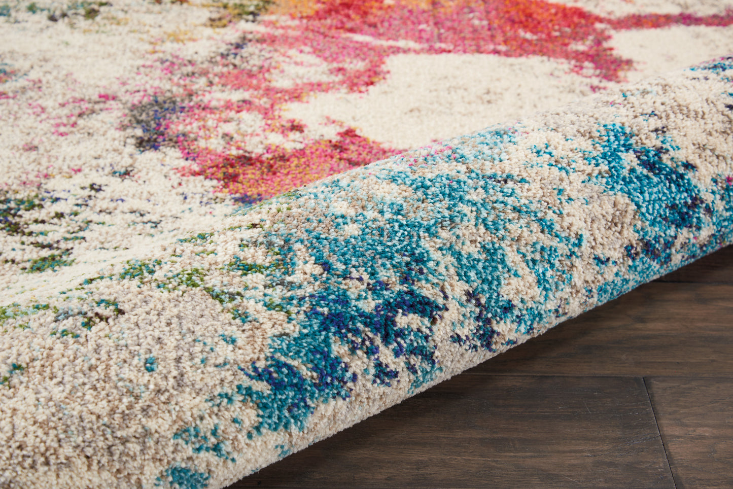 Colorful abstract shag area rug with beige base, blue/teal accents and pink highlights, partially rolled on dark wood floor.
