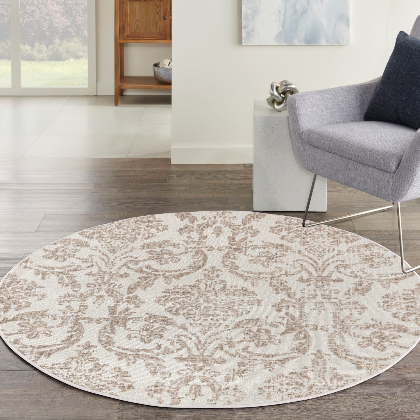 Round beige rug with taupe damask pattern on dark wood floor beside a gray armchair in a bright living room.