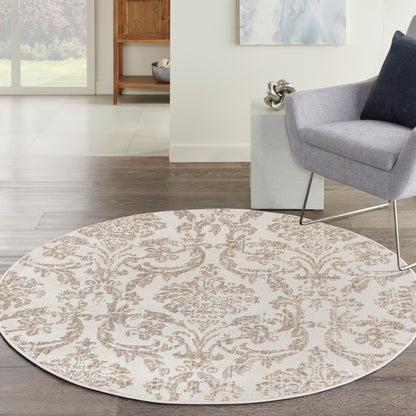 Round beige rug with taupe damask pattern on dark wood floor beside a gray armchair in a bright living room.